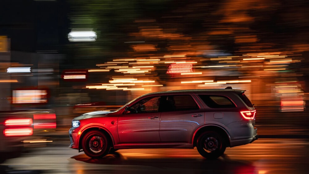 "Esta imagen muestra un Dodge Durango de color plateado conduciendo de noche con un fondo borroso, lo que indica movimiento. Las luces delanteras y traseras del vehículo están encendidas, y las luces circundantes crean un efecto dinámico, enfatizando la velocidad y el movimiento del coche. El vehículo está equipado con un motor HEMI V8, conocido por su potencia y rendimiento. Esta imagen es interesante porque captura la esencia del movimiento y la velocidad en un entorno urbano, destacando el rendimiento del vehículo y la vibrante vida nocturna de la ciudad.