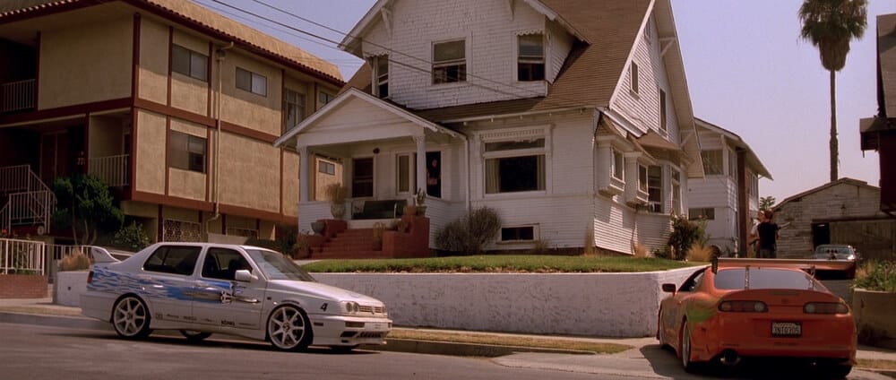La imagen muestra la casa de Dominic Toretto de la primera película de "Fast and Furious". Frente a la casa, hay dos coches icónicos estacionados: un Toyota Supra naranja con un gran alerón trasero y un Volkswagen Jetta blanco con calcomanías azules y ruedas de posventa. La casa tiene un porche con escalones que conducen a la entrada y está situada junto a un edificio de apartamentos más grande. Esta escena es emblemática para los fanáticos de la franquicia, ya que captura un momento clave de la película.