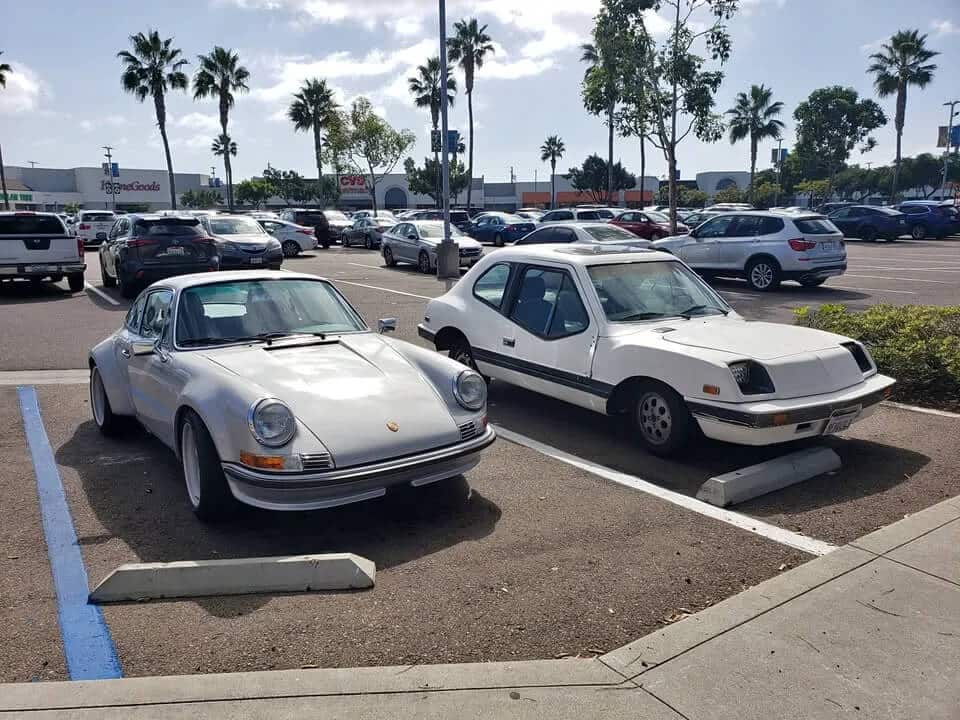 Imagen del Electrek UNCAR, un coche eléctrico único de 1979 fabricado por Unique Mobility, estacionado junto a un Porsche 911. El Electrek UNCAR es blanco con un diseño distintivo, incluyendo una parte trasera inclinada y un frente cuadrado. El Porsche 911, conocido por su diseño deportivo y elegante, está estacionado al lado del Electrek, creando un contraste interesante entre los dos vehículos. El fondo muestra un paisaje escénico con árboles y un cuerpo de agua, añadiendo un toque de serenidad a la escena.