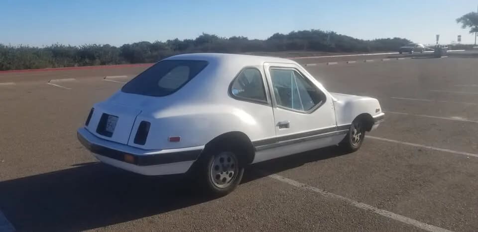 Imagen del Electrek UNCAR, un coche eléctrico único de 1979 fabricado por Unique Mobility. El coche es blanco con un diseño distintivo, incluyendo una parte trasera inclinada y un frente cuadrado. Está estacionado en un aparcamiento vacío, destacando su diseño inusual y llamativo. Este vehículo es conocido por su motor de 32 caballos de fuerza de General Electric y su transmisión manual de 4 velocidades de Volkswagen Fox. El Electrek UNCAR es un modelo raro y poco común, lo que lo hace interesante para los entusiastas de los automóviles eléctricos y coleccionistas.