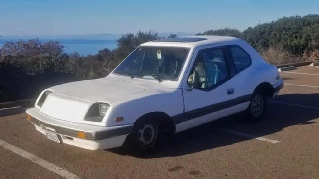 Imagen del Electrek UNCAR, un coche eléctrico único de 1979 fabricado por Unique Mobility. El coche es blanco con un diseño distintivo, incluyendo una parte trasera inclinada y un frente cuadrado. Está estacionado en un aparcamiento con un fondo escénico de árboles y un cuerpo de agua. Este vehículo es conocido por su motor de 32 caballos de fuerza de General Electric y su transmisión manual de 4 velocidades de Volkswagen Fox. El Electrek UNCAR es un modelo raro y poco común, lo que lo hace interesante para los entusiastas de los automóviles eléctricos y coleccionistas.