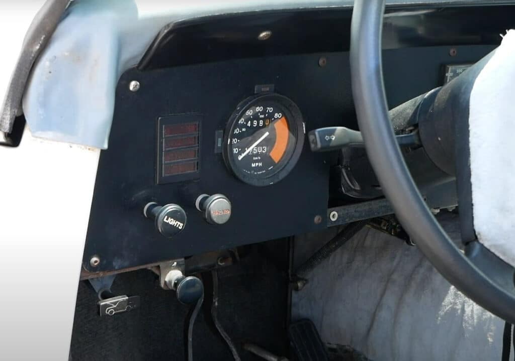 Imagen del tablero del Electrek UNCAR, un coche eléctrico único de 1979 fabricado por Unique Mobility. El tablero muestra el panel de instrumentos y los controles, incluyendo un velocímetro con una lectura máxima de 80 MPH, un odómetro que marca 49,810 millas y un cuentakilómetros parcial que marca 150.3 millas. Hay dos perillas etiquetadas como 'LIGHTS' y 'HEATER', y una palanca para los limpiaparabrisas. Además, hay un pequeño panel con un icono de un coche, probablemente indicando un control para el capó o el maletero.
