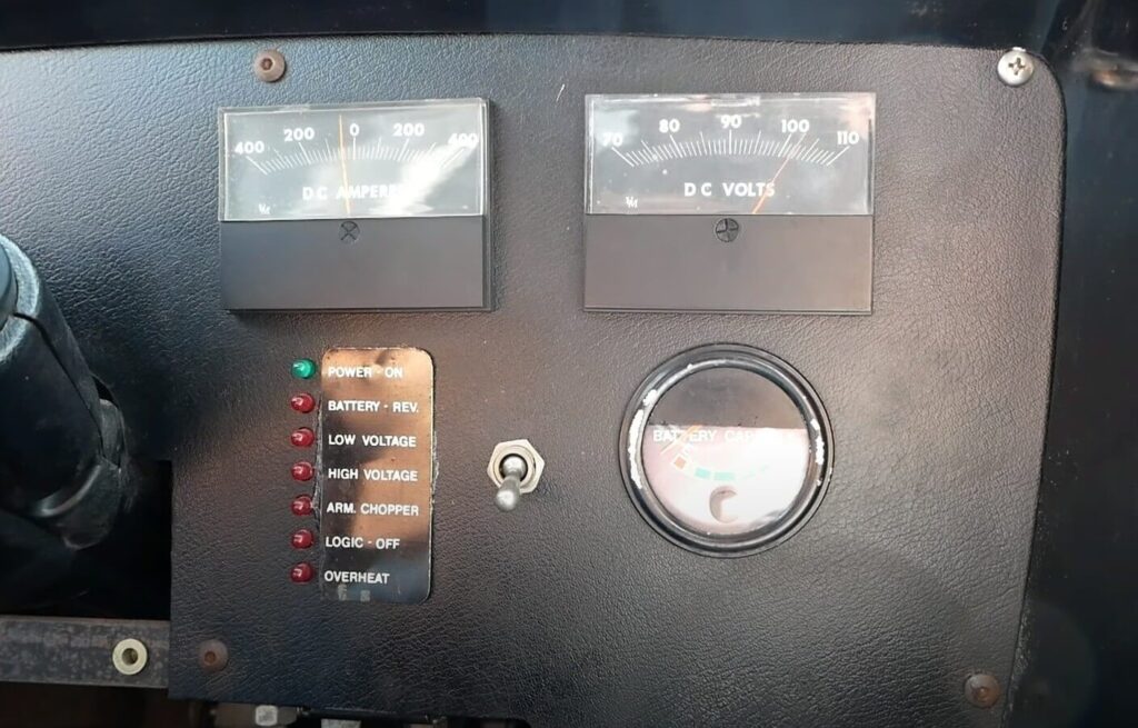 "Imagen del panel de control del Electrek UNCAR, un coche eléctrico único de 1979 fabricado por Unique Mobility. El panel muestra varios indicadores y medidores analógicos grandes en la parte superior: uno etiquetado como 'DC AMPERES' y otro como 'DC VOLTS'. Debajo de estos medidores, hay una serie de luces indicadoras con etiquetas como 'POWER ON', 'BATTERY - REV.', 'LOW VOLTAGE', 'HIGH VOLTAGE', 'ARM. CHOPPER', 'LOGIC - OFF' y 'OVERHEAT'. También hay un interruptor de palanca y un medidor circular etiquetado como 'BATTERY CAPACITY' con una escala de colores que indica diferentes niveles de carga de la batería.