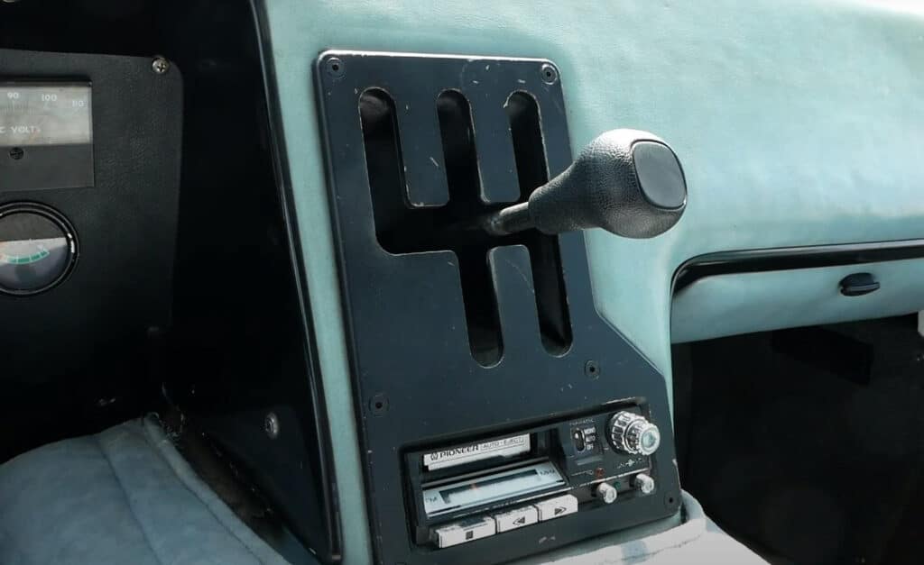 "Imagen del interior del Electrek UNCAR, un coche eléctrico único de 1979 fabricado por Unique Mobility. La imagen muestra la palanca de cambios y el área del tablero circundante. La palanca de cambios está posicionada en un selector de marchas con ranuras. Debajo de la palanca de cambios, hay un sistema de estéreo vintage de la marca Pioneer con reproductor de casetes. A la izquierda de la palanca de cambios, hay dos indicadores; uno parece ser un voltímetro y el otro es un indicador no identificado.