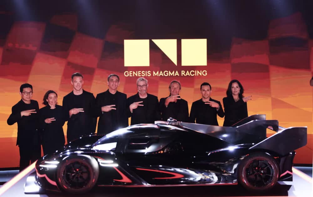 Un elegante coche de carreras negro Genesis GMR-001 se exhibe en un evento promocional. El fondo presenta el logotipo y el texto 'GENESIS MAGMA RACING'. El diseño aerodinámico y futurista del coche se destaca con una iluminación dramática, y un grupo de personas vestidas de negro están detrás, haciendo un gesto con la mano, posiblemente un símbolo del equipo o la marca.