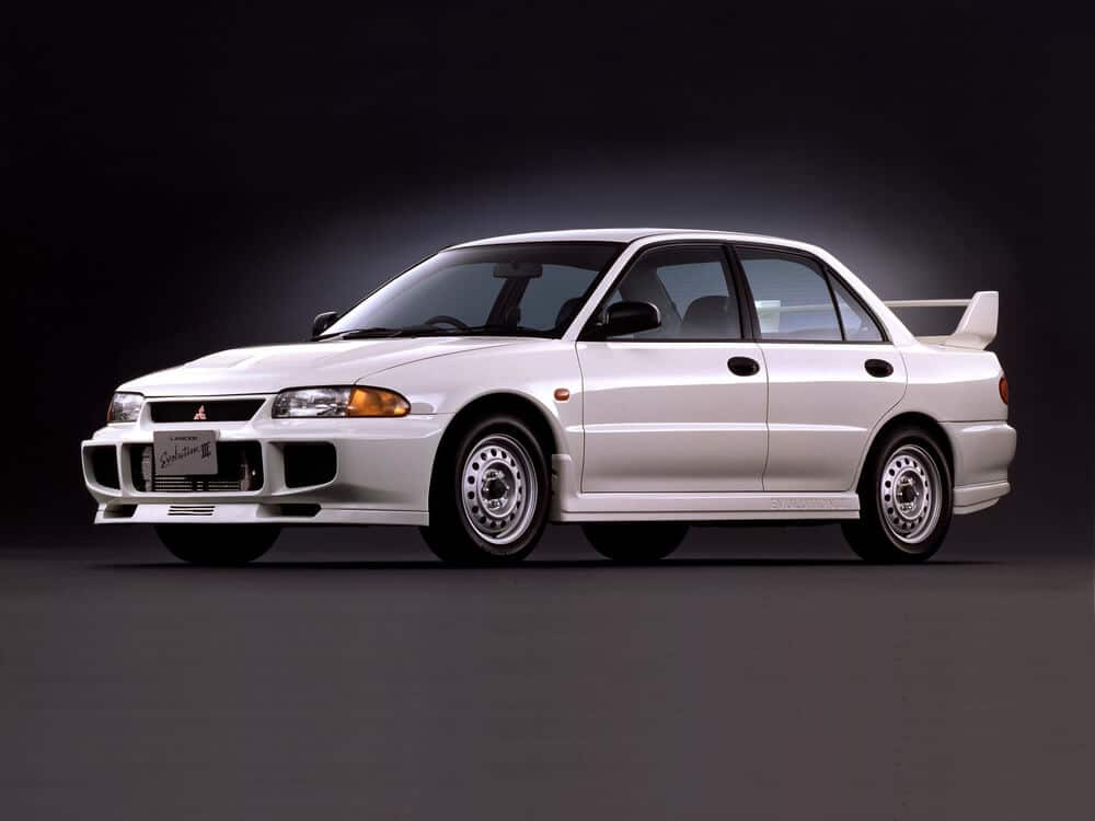 Un Mitsubishi Lancer Evolution III blanco, un sedán deportivo de alto rendimiento. Este coche es notable por su diseño agresivo, que incluye un parachoques delantero prominente con grandes tomas de aire, un alerón trasero y llantas de aleación deportivas. La serie Lancer Evolution es muy apreciada por su herencia en rally y su avanzado sistema de tracción en las cuatro ruedas, lo que lo convierte en una opción popular entre los entusiastas de los automóviles y los fanáticos del automovilismo.