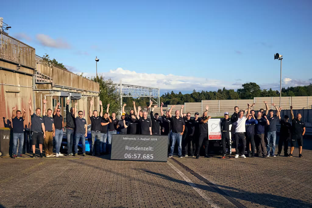 La imagen muestra un grupo de personas celebrando al aire libre junto a un edificio y una pista de carreras. Todos tienen los brazos levantados en señal de celebración. En primer plano, hay un cartel con el siguiente texto: "Mittwoch, 7. August 2019. Rundenzeit: 06:57.685." Esta imagen es interesante y relevante porque muestra el tiempo de vuelta del Ford Mustang GTD en el circuito de Nürburgring, destacando un logro significativo en el mundo del automovilismo.