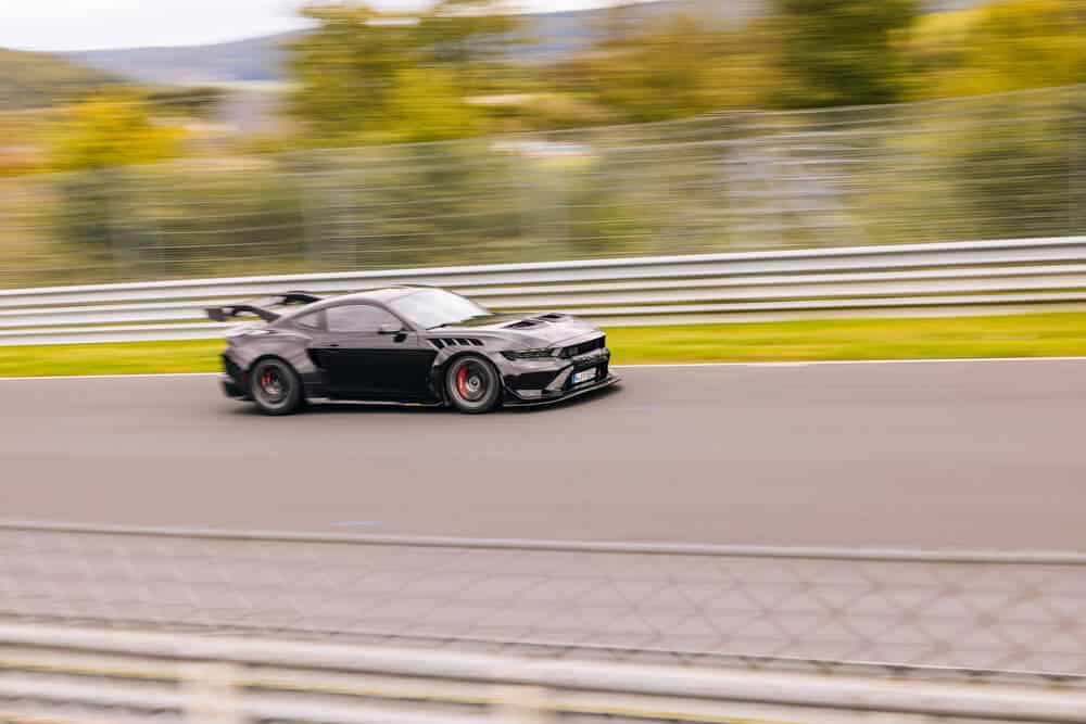 La imagen muestra un Ford Mustang GTD negro corriendo en el circuito de Nürburgring. El coche está capturado en movimiento, enfatizando su velocidad y capacidades de rendimiento. El fondo está ligeramente borroso, destacando el movimiento del coche. Esta imagen es interesante y relevante porque muestra un coche de alto rendimiento en uno de los circuitos de carreras más desafiantes y renombrados del mundo.