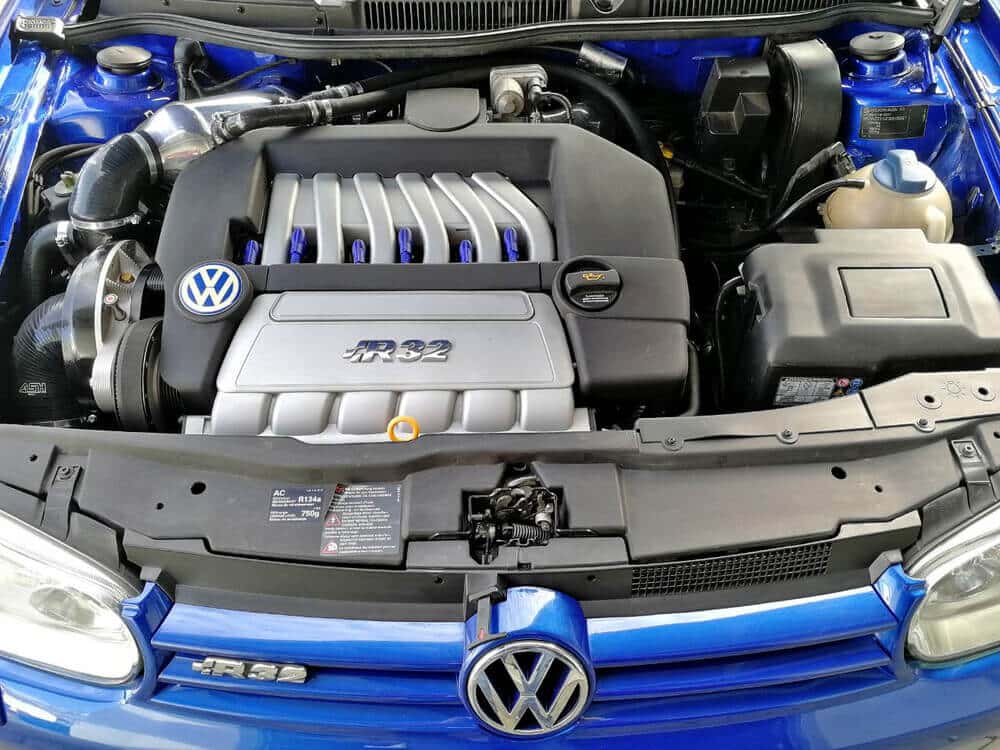 Esta imagen muestra el compartimento del motor de un vehículo Volkswagen, específicamente un modelo R32. La cubierta del motor exhibe prominentemente la insignia 'R32', indicando que se trata de una versión de alto rendimiento del Volkswagen Golf. El compartimento del motor está limpio y bien organizado, con componentes visibles como el sistema de admisión de aire, la batería y varios depósitos de fluidos. El motor VR6, conocido por su configuración en V estrecha con seis cilindros, es el corazón de este vehículo, proporcionando un equilibrio entre potencia y eficiencia. El logotipo de Volkswagen también se muestra de manera destacada en la cubierta del motor. Esta imagen es interesante y relevante porque destaca la ingeniería y el diseño de un vehículo orientado al rendimiento, mostrando la atención al detalle y la marca que Volkswagen incorpora en sus modelos de alto rendimiento.