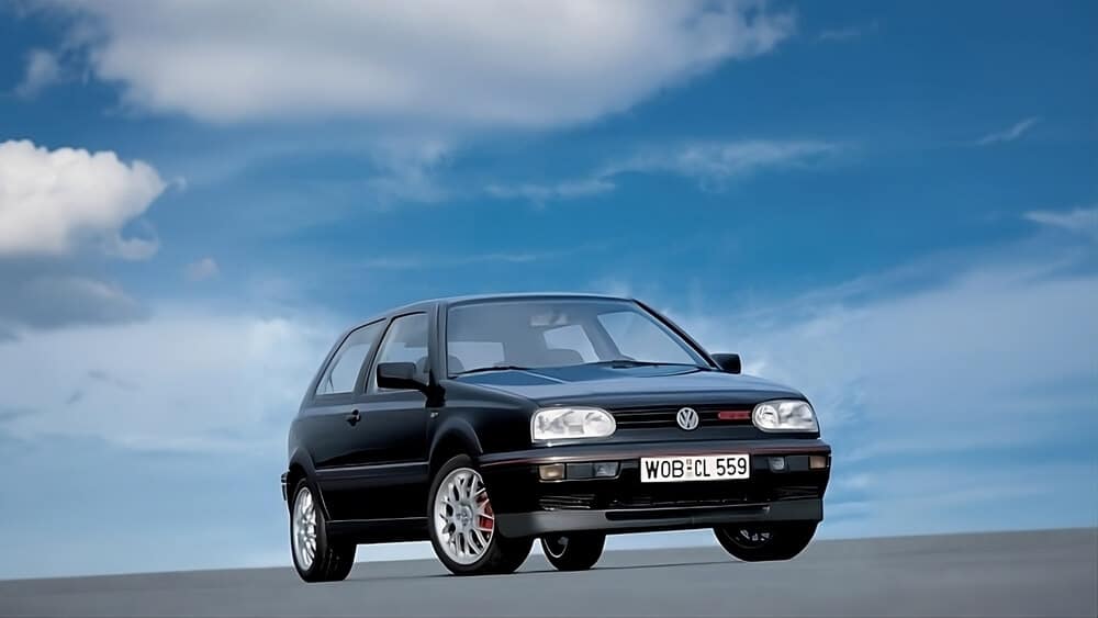"Esta imagen muestra un automóvil Volkswagen Golf VR6 de color negro, estacionado sobre una superficie plana con un cielo azul y algunas nubes en el fondo. El automóvil tiene una matrícula que dice 'WOB CL 559'. El vehículo es un modelo de tres puertas y se destaca por su diseño clásico y deportivo, característico de los modelos VR6.