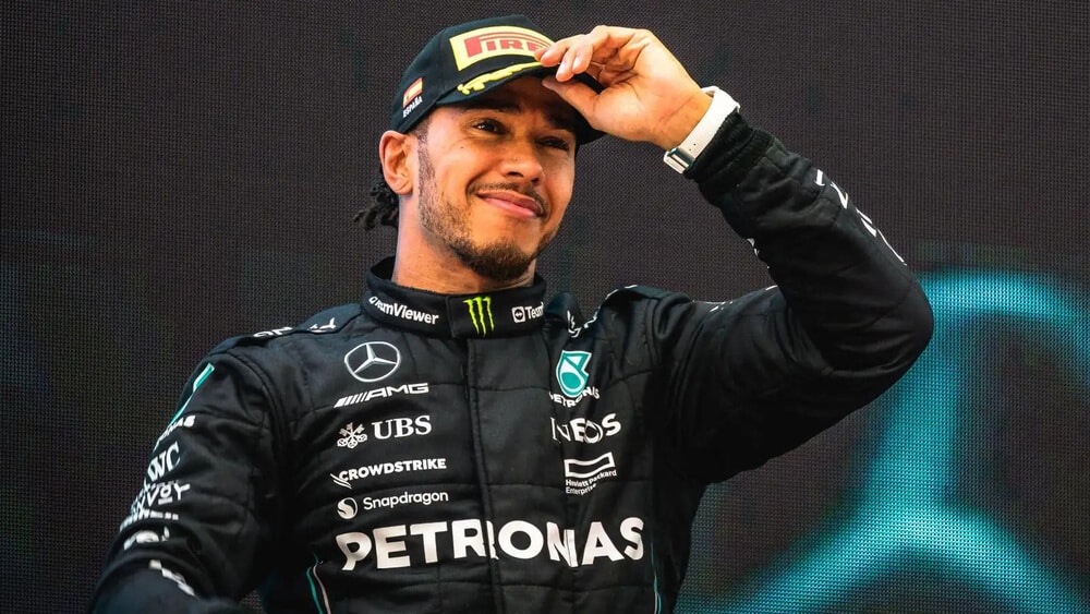 La imagen muestra a Lewis Hamilton, piloto de Fórmula 1, con un traje de carreras negro que lleva varios logotipos de patrocinadores, incluyendo Mercedes-Benz, Petronas y Monster Energy. Hamilton está sonriendo y ajustando su gorra negra con el logotipo de Pirelli. El fondo muestra una pantalla o banner grande con un logotipo borroso. Esta imagen es interesante y relevante ya que captura un momento de celebración o satisfacción de Hamilton, destacando su éxito y carisma en el mundo del automovilismo.