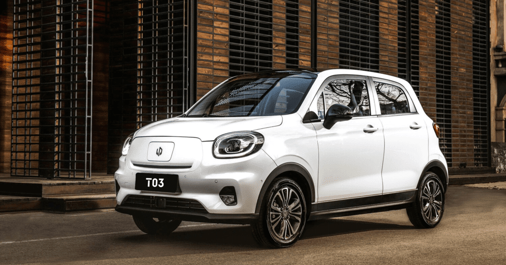 Imagen de un Leapmotor T03 blanco, un vehículo eléctrico compacto, estacionado en un entorno urbano. El coche está posicionado en un ángulo que muestra su perfil frontal y lateral. El T03 presenta un diseño moderno con faros redondeados, una carrocería elegante y detalles en negro en las ruedas y la parte inferior del vehículo. Al fondo, se puede ver un edificio con un diseño contemporáneo, con paneles de madera y rejillas metálicas, que complementa la estética moderna del coche. Esta imagen es relevante ya que destaca el diseño y la idoneidad urbana del Leapmotor T03, un vehículo eléctrico que representa avances en el transporte sostenible.