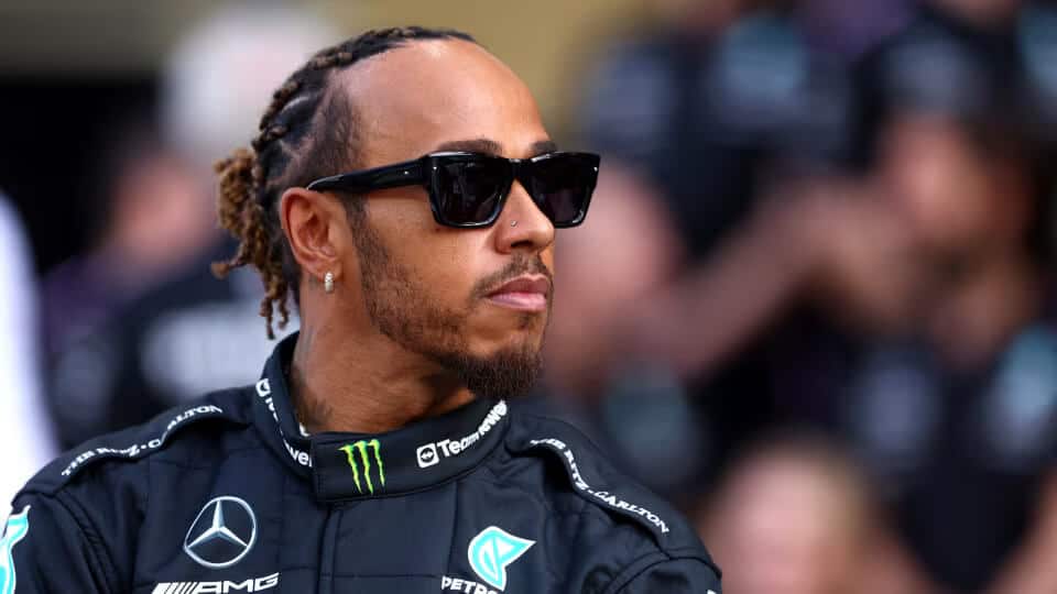 La imagen muestra a Lewis Hamilton, piloto de Fórmula 1, vistiendo un traje de carreras negro con varios logotipos de patrocinadores, incluyendo Mercedes-Benz, Petronas, Monster Energy y AMG. Hamilton tiene el cabello trenzado y lleva gafas de sol. El fondo está desenfocado, pero parece ser un evento de carreras o un entorno similar. Esta imagen es interesante y relevante porque muestra a un piloto profesional en el contexto de la Fórmula 1, destacando su asociación con el equipo Mercedes-Benz y sus patrocinadores.