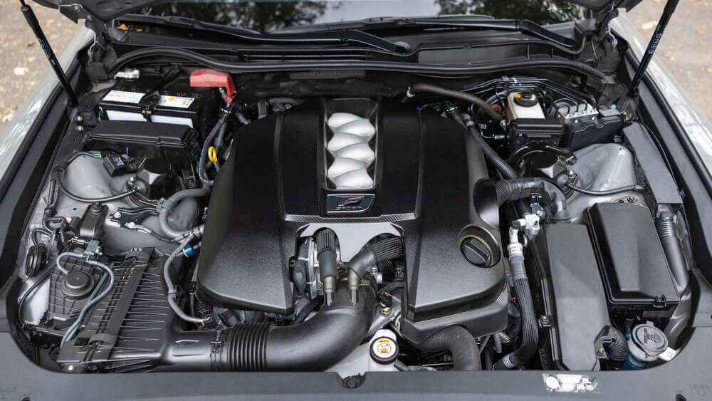 La imagen muestra el compartimento del motor de un Lexus IS500. El motor es una característica prominente, con una gran cubierta negra que tiene el logotipo "F" en el centro, indicando que es parte de la línea de rendimiento Lexus F. El compartimento del motor está bien organizado, con varios componentes como la batería, la admisión de aire y los depósitos de fluidos claramente visibles. Esta imagen es interesante y relevante porque destaca el potente motor V8, que es un punto clave de venta del IS500, mostrando su diseño y ingeniería orientados al rendimiento.