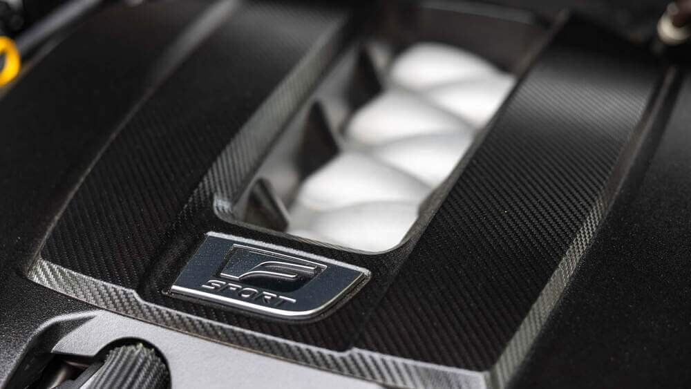 La imagen muestra una vista detallada de la cubierta del motor de un Lexus IS500 con un acabado de fibra de carbono. La cubierta presenta prominentemente la insignia "F SPORT", indicando que este es un modelo orientado al rendimiento de Lexus. El material de fibra de carbono y la marca "F SPORT" sugieren un enfoque en el alto rendimiento y la estética deportiva, lo cual es interesante y relevante para los entusiastas de los coches y aquellos interesados en el diseño y la ingeniería automotriz.
