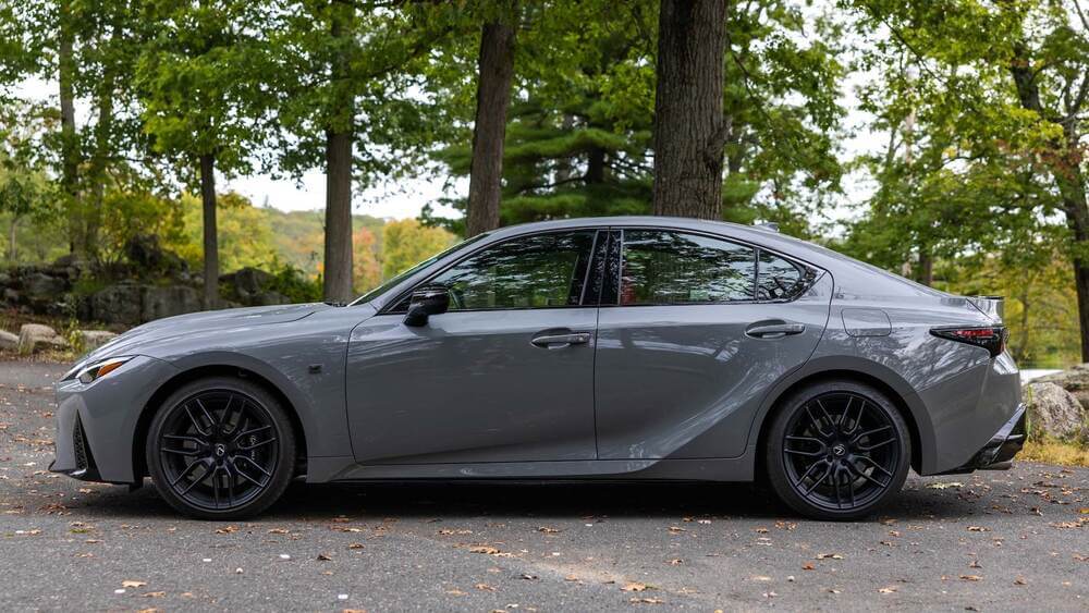 La imagen muestra una vista lateral de un Lexus IS500, un sedán deportivo de lujo, estacionado sobre una superficie pavimentada en un área boscosa. El coche tiene un diseño elegante y moderno, con llantas de aleación negras, una carrocería aerodinámica y ventanas tintadas. El entorno natural con árboles y follaje verde crea un contraste atractivo con el coche, destacando su estética y atractivo visual. Esta imagen es interesante y relevante para los entusiastas de los coches y aquellos interesados en vehículos de lujo y alto rendimiento.