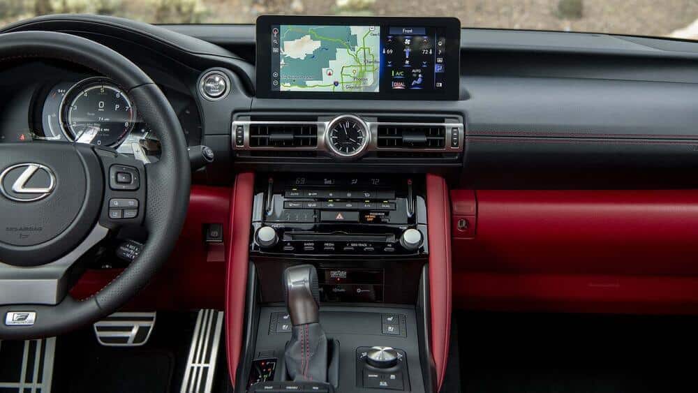 La imagen muestra el interior de un Lexus, enfocándose en el tablero y la consola central. El volante, el cuadro de instrumentos y el sistema de infoentretenimiento son los elementos más destacados. La pantalla del sistema de infoentretenimiento muestra un mapa de navegación e información sobre el control del clima. El tablero combina cuero negro y rojo, con varios botones y perillas para controlar el audio, el clima y otras funciones del vehículo. El diseño es elegante y moderno, resaltando el lujo y la tecnología avanzada del vehículo. Esta imagen es interesante y relevante ya que muestra las características de alta gama y el atractivo estético del interior del Lexus.