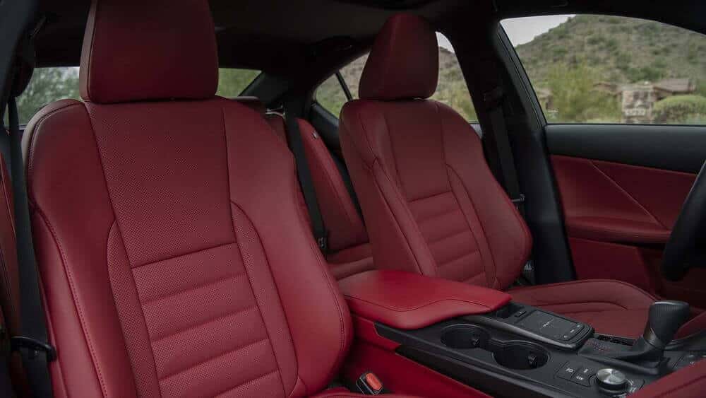 La imagen muestra el interior de un coche, enfocándose en los asientos delanteros y la consola central. Los asientos están tapizados en cuero rojo, con un diseño que incluye costuras detalladas y perforaciones para ventilación. La consola central cuenta con un reposabrazos acolchado, portavasos y varios controles, incluyendo un selector de marchas y un dial giratorio. El fondo fuera de la ventana del coche muestra un paisaje natural con colinas y algunos edificios. Esta imagen es interesante ya que destaca el diseño lujoso y elegante del interior del coche, lo cual puede atraer a posibles compradores interesados en la comodidad y la estética de un vehículo.