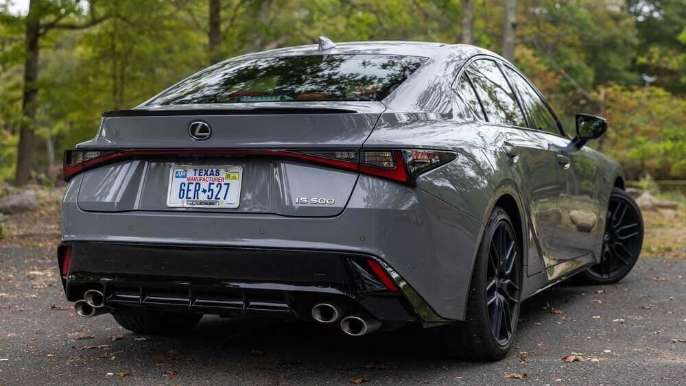 La imagen muestra la vista trasera de un Lexus IS500 gris estacionado en una carretera con un fondo boscoso. El coche tiene una matrícula de fabricante de Texas con el número "6ER 527". El vehículo presenta un diseño elegante con luces traseras distintivas, un alerón trasero y cuatro tubos de escape, lo que indica que es un modelo orientado al rendimiento. La insignia "IS 500" es visible en el maletero, lo que identifica el modelo del coche. Esta imagen es interesante porque destaca el diseño y las características de un sedán de lujo de alto rendimiento, lo cual puede atraer a los entusiastas de los coches y a posibles compradores.