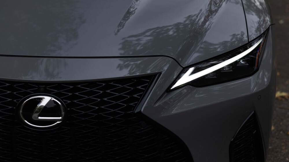 La imagen muestra la parte frontal de un vehículo Lexus, enfocándose en la parrilla y el faro izquierdo. El logotipo de Lexus se exhibe de manera prominente en el centro de la parrilla, que tiene un patrón de malla distintivo. El faro presenta un diseño moderno y elegante con elementos LED. El capó del coche y parte del parachoques delantero también son visibles, reflejando el entorno. Esta imagen es interesante y relevante ya que destaca los elementos de diseño y la marca del Lexus IS500, mostrando su lujo y atención al detalle.