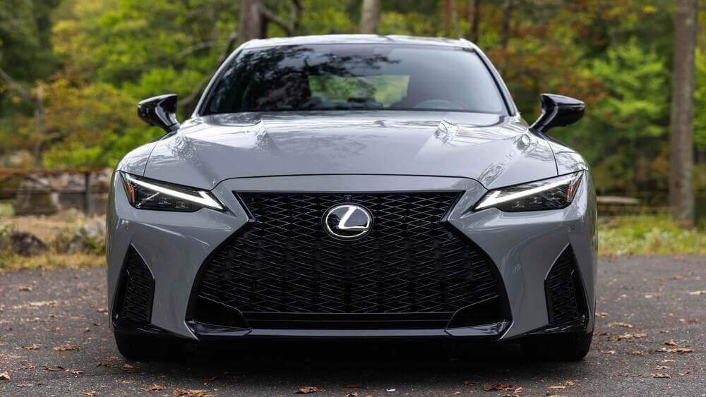 La imagen muestra la vista frontal de un Lexus IS500 gris estacionado en una carretera con un fondo boscoso. El coche tiene una parrilla negra prominente con un patrón de malla, faros LED elegantes y un diseño aerodinámico. El logotipo de Lexus está centrado en la parrilla. El entorno natural con árboles y vegetación sugiere que el coche está estacionado al aire libre. Esta imagen es interesante ya que destaca los elementos de lujo y diseño del Lexus, mostrando su atractivo estético y características modernas.