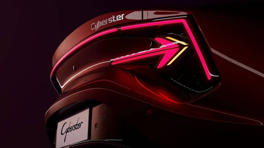 El extremo trasero de un MG Cyberster con el nombre 'Cyberster' mostrado prominentemente en la parte trasera. El diseño presenta líneas modernas y elegantes con luces traseras angulares distintivas que tienen una apariencia futurista. El nombre del coche también se muestra en el área de la matrícula. La iluminación y los reflejos en la superficie del coche resaltan su acabado brillante y diseño aerodinámico.