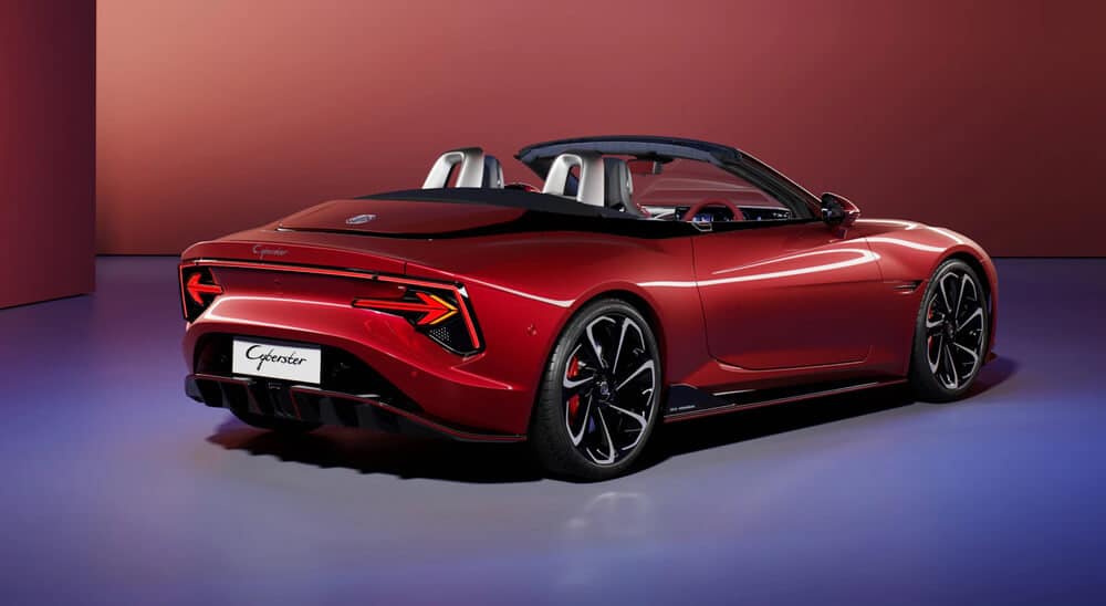 Un MG Cyberster rojo descapotable con un diseño elegante y moderno. El coche tiene luces traseras distintivas con un diseño angular único, grandes ruedas negras con pinzas de freno rojas y un difusor trasero negro. La matrícula trasera dice 'Cyberster'. El fondo es un degradado de tonos rojos y púrpuras, dando a la imagen una apariencia estilizada y futurista.
