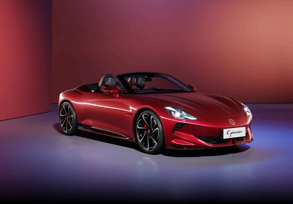 Un MG Cyberster rojo descapotable con un diseño elegante y moderno. El coche tiene luces traseras distintivas con un diseño angular único, grandes ruedas negras con pinzas de freno rojas y un difusor trasero negro. La matrícula trasera dice 'Cyberster'. El fondo es un degradado de tonos rojos y púrpuras, dando a la imagen una apariencia estilizada y futurista.