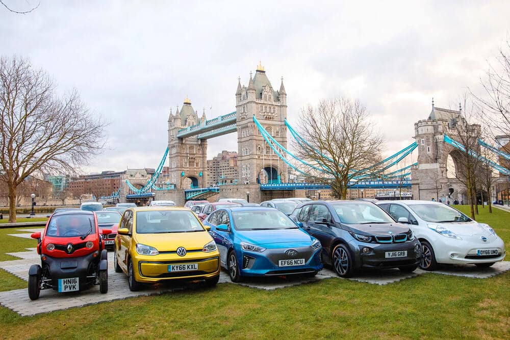 Una fila de varios coches eléctricos e híbridos estacionados frente al icónico Tower Bridge en Londres. Los autos incluyen un Renault Twizy rojo, un Volkswagen e-Up! amarillo, un Hyundai Ioniq azul, un BMW i3 negro y un Nissan Leaf blanco. El fondo del Tower Bridge añade un contexto cultural e histórico significativo a la imagen, destacando la yuxtaposición de vehículos eléctricos modernos con un monumento histórico. Esta imagen es interesante y relevante ya que muestra la creciente presencia e importancia de los vehículos eléctricos en entornos urbanos, promoviendo la sostenibilidad y la conciencia ambiental.