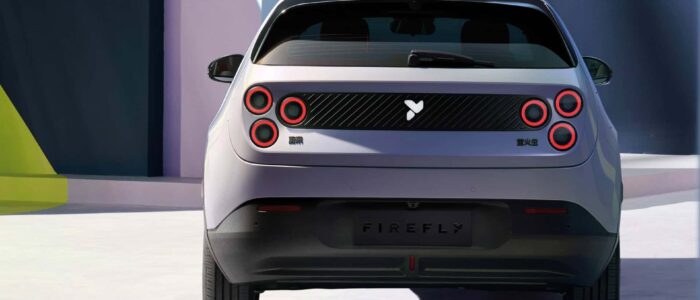 La parte trasera de un coche eléctrico NIO Firefly con un diseño moderno y elegante. El coche tiene cuatro luces traseras circulares, dos a cada lado, resaltadas con anillos rojos. Hay un logotipo en el centro de la parte trasera y texto en un alfabeto no latino en el lado izquierdo del panel trasero. El coche está situado en un entorno moderno y bien iluminado con formas geométricas y colores contrastantes en el fondo, sugiriendo un ambiente contemporáneo y estilizado. Esta imagen es interesante porque muestra el diseño y la marca de un nuevo o concepto de vehículo, destacando su atractivo estético y su posible posicionamiento en el mercado.