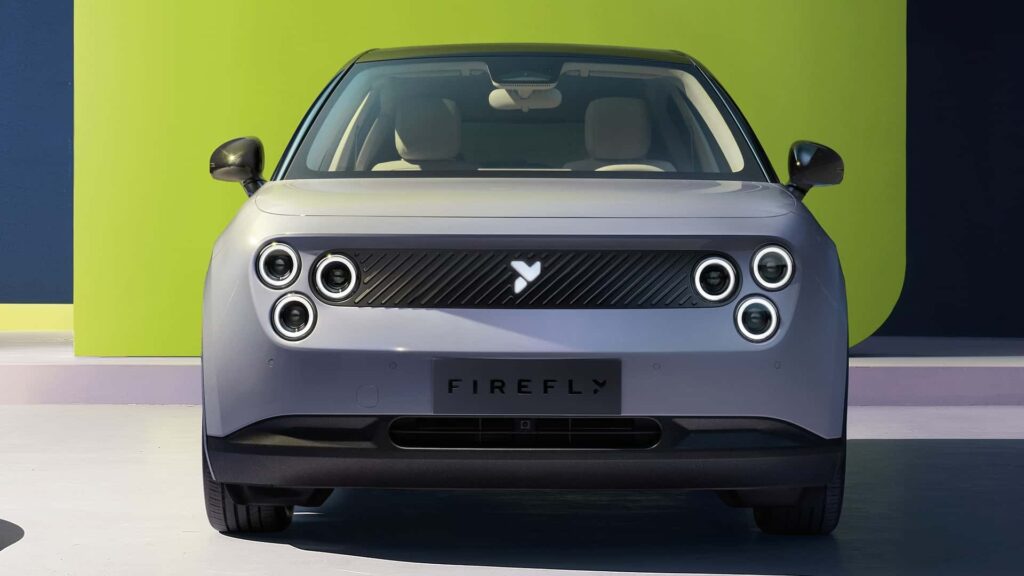 "Vista frontal de un coche eléctrico con la marca 'NIO Firefly' en el área de la matrícula. El coche tiene un diseño moderno y minimalista con cuatro faros circulares a cada lado y una parrilla elegante en el centro. El fondo presenta una combinación de colores verde y azul oscuro, añadiendo un contraste vibrante a la apariencia del coche. Esta imagen es interesante porque destaca los elementos de diseño únicos del NIO Firefly, que pueden atraer a aquellos interesados en el diseño automotriz y la innovación.