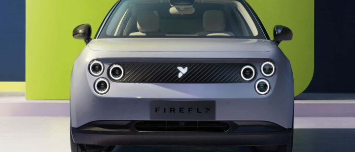 Vista frontal de un coche eléctrico con la marca 'NIO Firefly' en el área de la matrícula. El coche tiene un diseño moderno y minimalista con cuatro faros circulares a cada lado y una parrilla elegante en el centro. El fondo presenta una combinación de colores verde y azul oscuro, añadiendo un contraste vibrante a la apariencia del coche. Esta imagen es interesante porque destaca los elementos de diseño únicos del NIO Firefly, que pueden atraer a aquellos interesados en el diseño automotriz y la innovación.