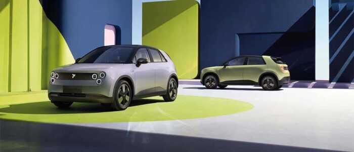 Dos vehículos eléctricos compactos NIO Firefly en un entorno estilizado y futurista. El coche en primer plano es de color gris claro con faros circulares distintivos y un diseño elegante. El coche en el fondo es de color verde y tiene un diseño similar. El escenario presenta grandes formas geométricas en colores vibrantes como verde y azul, creando un fondo visualmente impactante y moderno. Esta imagen es interesante y relevante ya que muestra el diseño y la estética de los nuevos vehículos eléctricos, destacando los avances en tecnología automotriz y transporte sostenible.