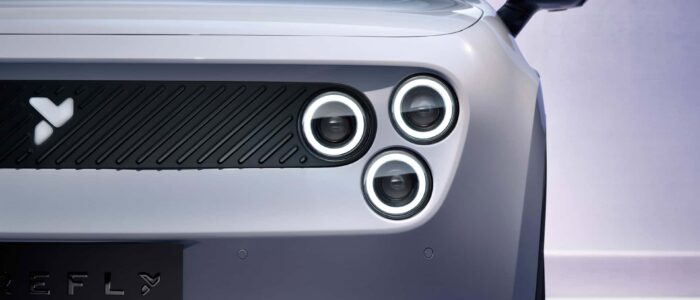 El frontal de un coche eléctrico moderno, centrado en el área de los faros. El coche presenta un diseño único con tres faros circulares a la derecha de la imagen, cada uno rodeado por anillos LED. A la izquierda de la imagen se ve una parrilla negra con líneas diagonales y un logotipo distintivo. Debajo de la parrilla, parte del nombre del coche, 'NIO Firefly', es visible, con un logotipo estilizado en forma de 'Y'. El diseño del coche es elegante y futurista, lo que lo hace interesante y relevante en el contexto de las tendencias de diseño automotriz moderno.