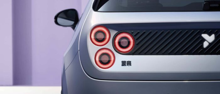 La parte trasera de un coche, centrada en las luces traseras y parte de la ventana trasera. El coche tiene un diseño distintivo con cuatro luces traseras circulares dispuestas en una línea vertical en el lado izquierdo. Hay un panel negro con líneas diagonales y un logotipo blanco en el lado derecho de las luces traseras. Debajo del panel negro, hay algunos caracteres, posiblemente en chino o japonés. La carrocería del coche está pintada en un color claro, y el fondo es una pared de color púrpura claro. Esta imagen es interesante porque destaca los elementos de diseño únicos y modernos del NIO Firefly, que pueden atraer a los entusiastas del automóvil o a posibles compradores.