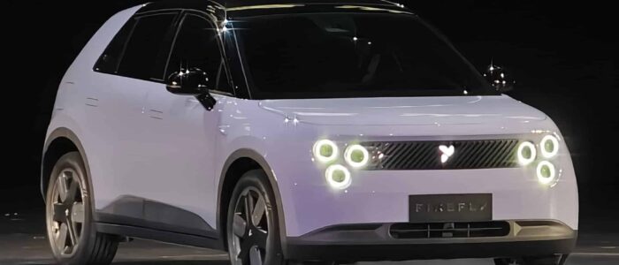 Un vehículo eléctrico compacto NIO Firefly con un diseño moderno y elegante. El coche tiene una carrocería blanca con detalles en negro, incluyendo el techo y los espejos laterales. En la parte frontal, se destacan los faros LED circulares dispuestos en pares a cada lado y una parrilla con un logotipo en forma de corazón en el centro. La matrícula tiene la palabra 'FIREFLY'. El coche está posicionado en un escenario o sala de exposición, sugiriendo que podría ser parte de un evento de presentación o lanzamiento. Esta imagen es interesante y relevante ya que muestra un nuevo modelo de vehículo eléctrico, destacando los avances en diseño y tecnología automotriz.
