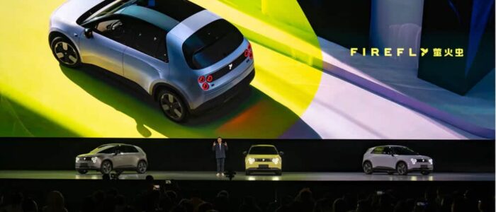 Un evento de presentación del NIO Firefly, un nuevo modelo de coche eléctrico. En el escenario hay tres coches físicos y una gran pantalla detrás de ellos muestra una imagen del NIO Firefly desde una perspectiva aérea. La pantalla también tiene el texto 'FIREFLY' junto con caracteres chinos. El evento parece estar muy concurrido, indicando un gran interés o importancia.