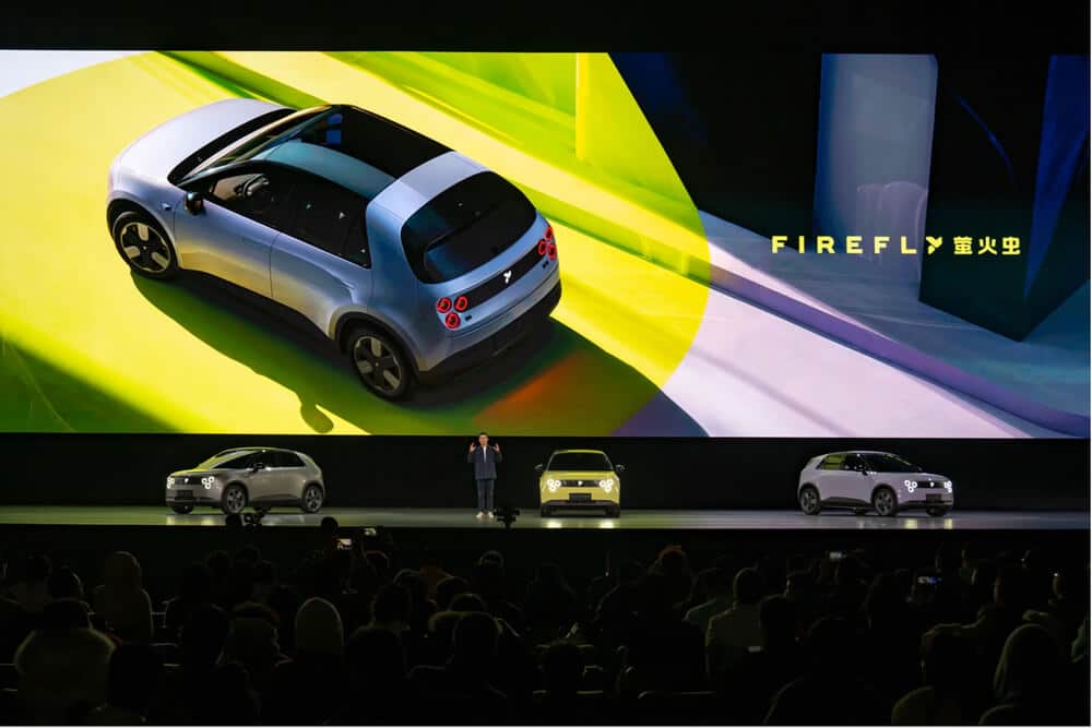 Un evento de presentación del NIO Firefly, un nuevo modelo de coche eléctrico. En el escenario hay tres coches físicos y una gran pantalla detrás de ellos muestra una imagen del NIO Firefly desde una perspectiva aérea. La pantalla también tiene el texto 'FIREFLY' junto con caracteres chinos. El evento parece estar muy concurrido, indicando un gran interés o importancia.