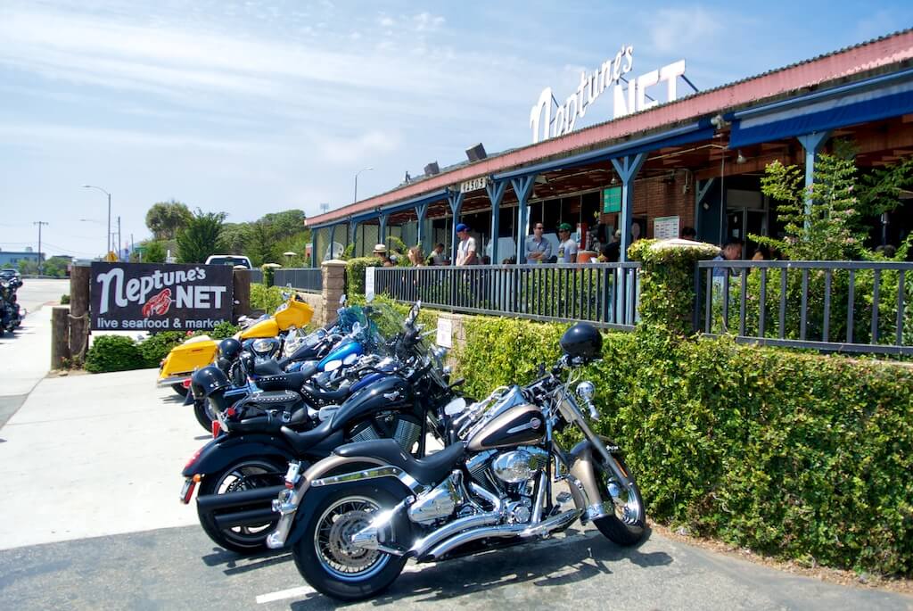 La imagen muestra el exterior de Neptune's Net, un conocido restaurante y mercado de mariscos. Varias motocicletas están estacionadas frente al restaurante, y se pueden ver personas en el área del patio. El cartel del restaurante se muestra de manera prominente, con el texto "Neptune's NET live seafood & market." Este lugar es notable por haber aparecido en la película "Fast and Furious 1," lo que lo convierte en un punto de interés para los fanáticos de la película.