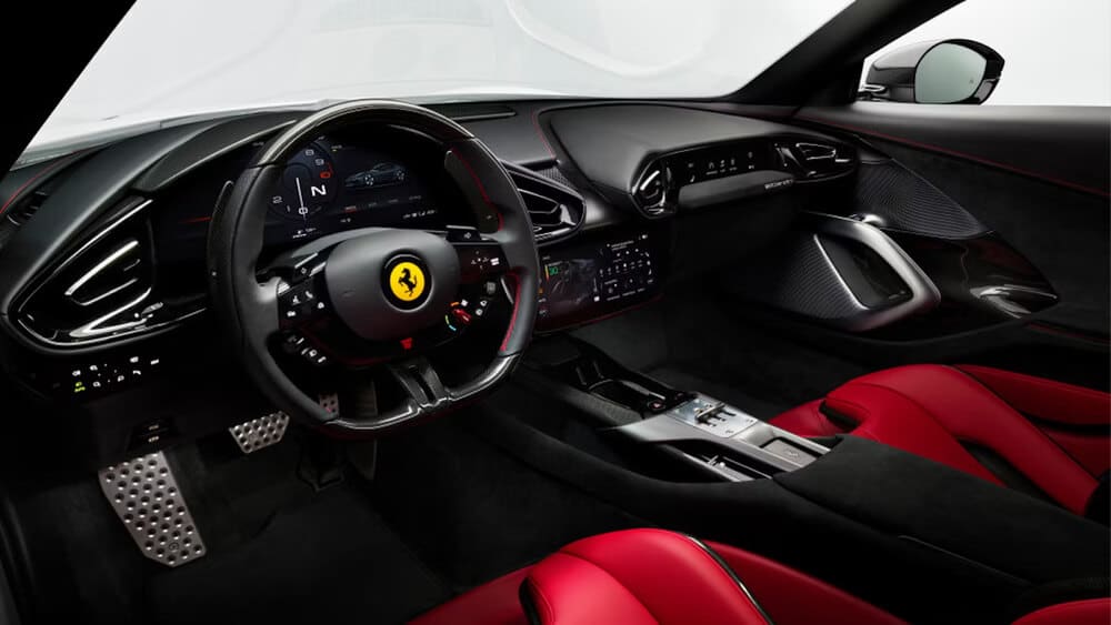 "La imagen muestra el interior de un coche deportivo de alta gama, específicamente el Ferrari 12 Cilindri. El volante presenta un logotipo prominente en el centro, rodeado de varios controles y botones. El salpicadero incluye una pantalla digital con un velocímetro, indicador de marchas y otra información del vehículo. El interior está diseñado con una combinación de materiales negros y rojos, incluyendo asientos de cuero y detalles en fibra de carbono. El diseño general es elegante y moderno, enfatizando el lujo y el rendimiento.