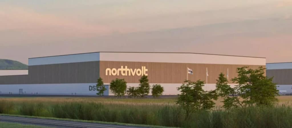 La imagen muestra un gran edificio industrial con el nombre "northvolt" destacado en un costado. El edificio está situado en una zona de césped con algunos árboles en primer plano. También hay algunas banderas visibles cerca del edificio. El cielo parece estar al amanecer o al atardecer, dando a la escena una luz cálida y suave. El diseño del edificio es moderno y limpio, lo que sugiere que es una instalación de una empresa contemporánea, probablemente involucrada en tecnología o manufactura.