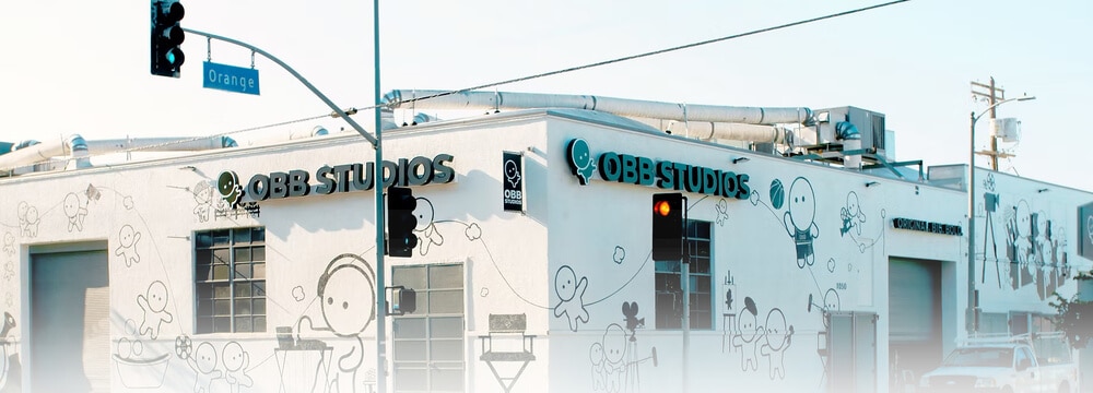 La imagen muestra un edificio con el nombre "OBB STUDIOS" destacado en la fachada. El edificio está decorado con varios dibujos de personajes de estilo caricaturesco involucrados en diferentes actividades. Hay una señal de calle que indica la intersección de Orange Street. El edificio parece ser un estudio de cine, como sugiere el nombre y las decoraciones artísticas. Esta imagen es interesante porque revela la ubicación real de lo que se conoce como Racer's Edge en la franquicia de "Fast and Furious", que en realidad es un estudio de cine.