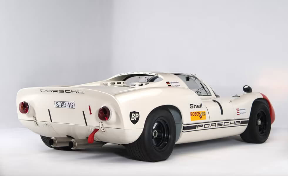 Vista trasera izquierda de un coche de carreras clásico, específicamente un Porsche, con varios adhesivos de patrocinadores, incluyendo Shell, Bosch y BP. La matrícula dice 'S-XR 46'. El coche tiene un diseño aerodinámico distintivo con un perfil bajo, grandes neumáticos traseros y tubos de escape dobles. Las luces traseras son redondas y rojas, y hay una flecha roja apuntando hacia abajo cerca de la luz trasera izquierda. El coche también tiene ruedas negras y una franja negra a lo largo del costado con la palabra 'Porsche' escrita en ella.