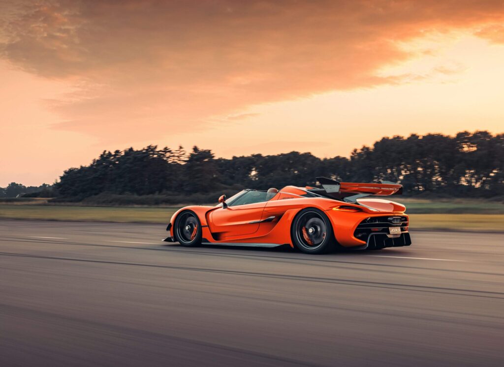 La imagen muestra un Koenigsegg Jesko naranja, un hipercoche de alto rendimiento, conduciendo a alta velocidad en una carretera o pista. El coche está capturado en movimiento con un fondo borroso de árboles y un cielo al atardecer, lo que enfatiza su velocidad y diseño dinámico. El Jesko es conocido por su ingeniería avanzada, motor potente y características aerodinámicas, lo que lo convierte en un tema significativo e interesante en el mundo automotriz.