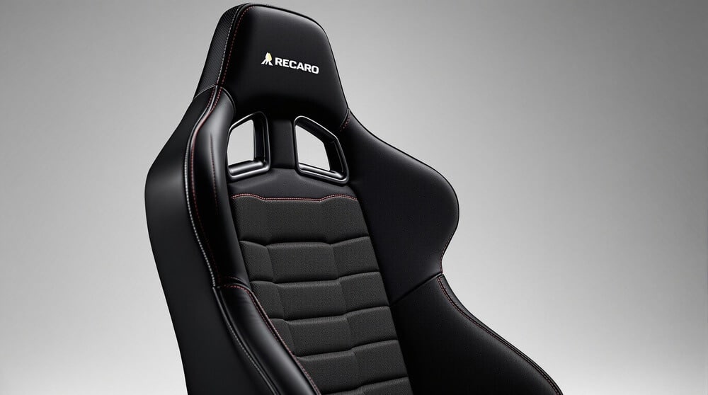 La imagen muestra un asiento de carreras de alto rendimiento, específicamente un asiento de la marca Recaro. El asiento está diseñado con contornos ergonómicos y presenta una combinación de materiales de cuero y tela. Es de color negro con costuras rojas y tiene ranuras para los arneses de seguridad. El logotipo de Recaro se muestra de manera prominente en la parte superior del asiento. Este tipo de asiento es interesante y relevante para los entusiastas del automovilismo y profesionales que buscan un mayor soporte y seguridad durante la conducción a alta velocidad o en condiciones de carrera.