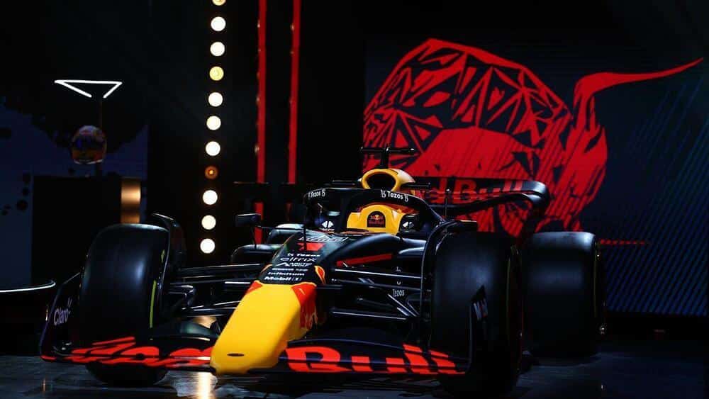 "Un coche de Fórmula 1 del equipo Red Bull Racing, con un llamativo esquema de colores negro y rojo, mostrando el logotipo de Red Bull en el morro y los laterales. El fondo incluye un gran gráfico estilizado de un toro rojo, que forma parte de la marca del equipo. El coche está iluminado por focos, destacando su diseño aerodinámico y elegante. Esta imagen es interesante y relevante ya que muestra la ingeniería avanzada y el diseño de un vehículo de alto rendimiento, que es un aspecto clave de las carreras de Fórmula 1.