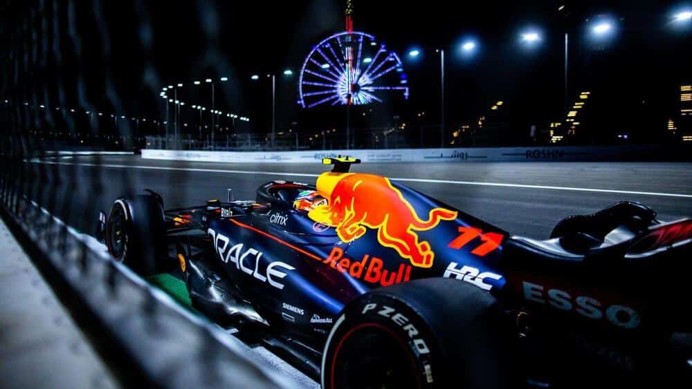 Un coche de Fórmula 1 de Red Bull Racing en un circuito nocturno. El coche está decorado con los logotipos de 'Oracle', 'Red Bull' y otros patrocinadores como 'HRC', 'ESSO' y 'Pirelli'. El número '11' es visible en el coche. En el fondo, se ve una noria iluminada y varias farolas que iluminan la pista. La imagen captura la naturaleza dinámica y de alta velocidad de las carreras de Fórmula 1, destacando el diseño del coche y el entorno de carreras nocturnas.