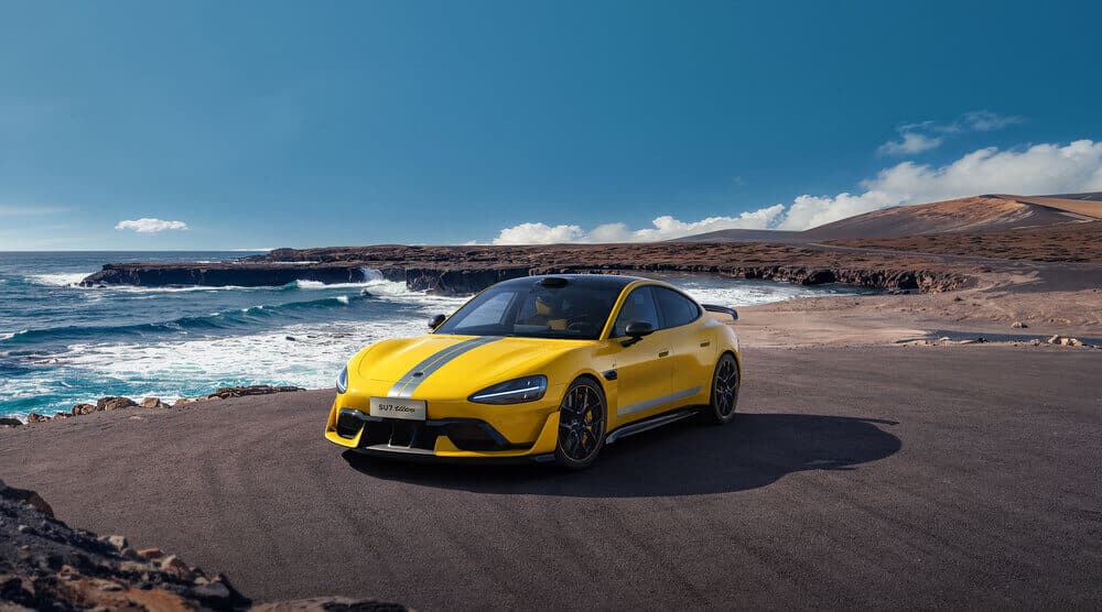 La imagen muestra un Xiaomi SU7 Ultra, un coche deportivo amarillo con una franja de carreras gris, estacionado en una zona pavimentada cerca de un paisaje costero. El coche está en primer plano, con las olas del océano rompiendo contra los acantilados rocosos en el fondo. El cielo está despejado con algunas nubes, y el entorno general parece ser una área costera remota y pintoresca. La matrícula del coche dice "SU7 Ultra." Esta imagen es interesante porque combina el diseño moderno y elegante del coche deportivo con la belleza natural del entorno costero, creando un contraste llamativo entre la tecnología y la naturaleza.