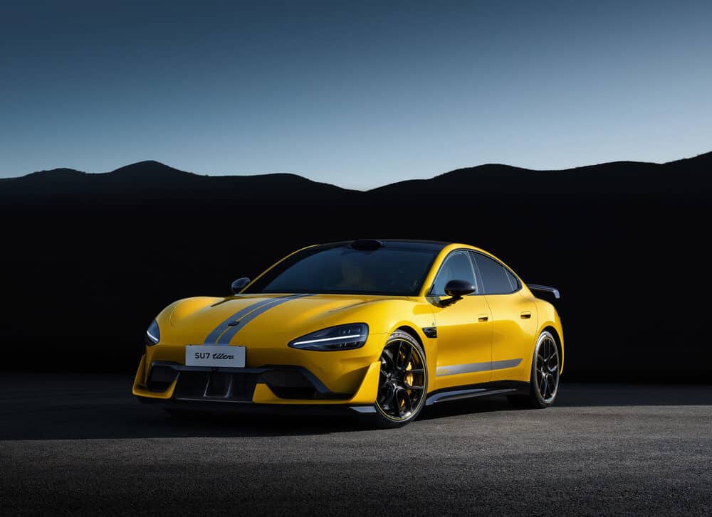 La imagen muestra un Xiaomi SU7 Ultra, un coche deportivo amarillo con acentos negros y franjas de carreras. El coche está posicionado en una carretera con un paisaje montañoso de fondo, bajo un cielo despejado. El diseño elegante, el color vibrante y las características deportivas hacen que este coche sea visualmente impactante y relevante para los entusiastas del automovilismo o posibles compradores interesados en vehículos de alto rendimiento.