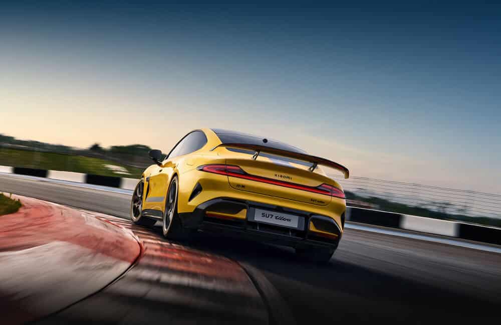 La imagen muestra un Xiaomi SU7 Ultra, un coche deportivo amarillo con acentos negros y franjas de carreras, conduciendo en una pista de carreras. La vista es desde la parte trasera, destacando su diseño aerodinámico y elegante, con el nombre de la marca "Xiaomi" en la parte trasera. La pista de carreras tiene bordillos rojos y blancos, y el fondo muestra un cielo despejado con un toque de atardecer, añadiendo una sensación dinámica y de alta velocidad a la escena. Esta imagen es interesante y relevante ya que muestra un vehículo de alto rendimiento en acción, enfatizando la velocidad, el diseño y la ingeniería del coche.